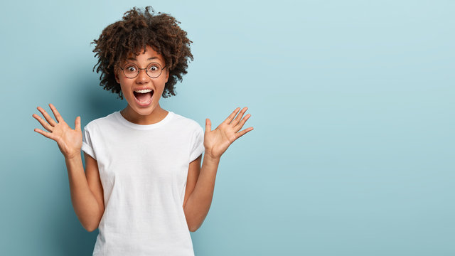 Curly optimistic woman raises palms from joy, happy to receive awesome present from someone, shouts loudly, dressed in casual white t shirt, isolated on blue background. Excited Afro female yells