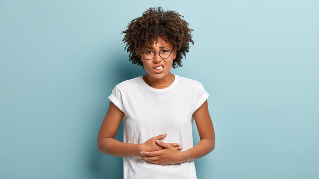 Dissatisfied young woman has food poisoning, keeps hands on belly, suffers from menstrual period cramp, feels discomfort in stomach, wears casual clothes, frowns face, isolated on blue background