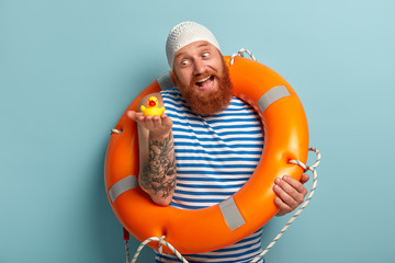 Playful joyous man with red thick beard, holds small toy duckling, has fun on shore, enjoys safe swimming with lifebuoy and swimhat, spends summer holidays, isolated on blue background. Recreation
