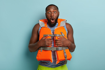Stupefied dark skinned man dressed in life jacket, shocked to have emergency situation, has thick...