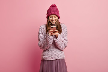 Amused upbeat pleased young woman holds smartphone, wears winter hat and knitted jumper, poses against pink background, reads funny story on social media. People, modern technologies concept
