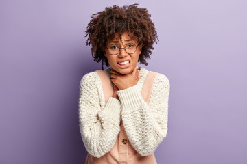 Displeased dark skinned woman suffers from suffocation, keeps both hands on throat, clenches teeth, has curly Afro hair, wears white knitted jumper, isolated over purple background, has asthma - obrazy, fototapety, plakaty