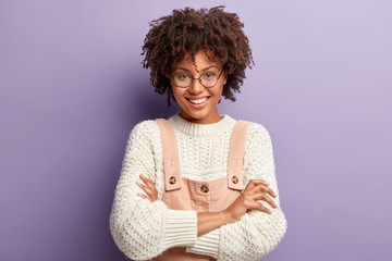 Studio shot of pleasant looking friendly woman with Afro hairstyle, enjoys life, smiles gently, wears round spectalces, white sweater and pink overalls, feels happy and upbeat. Pleasant emotions
