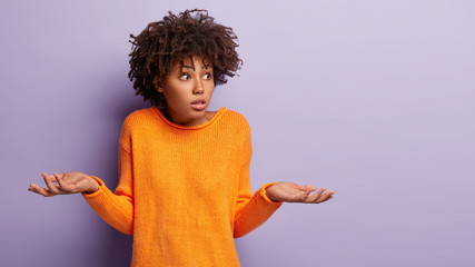 Insecure puzzled dark skinned female has Afro hairstyle, spreads hands with doubt, dressed in...