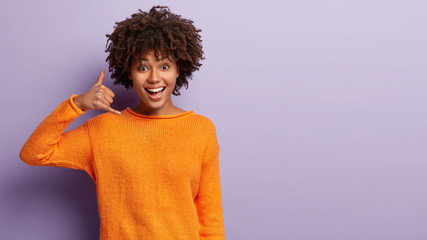 Delighted young female model with happy expression, makes call gesture, asks being in touch, wears casual orange jumper, isolated over purple background with free space for your advertisement