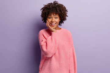 Pleased dark skinned woman has sincere smile, reacts on funny story, touches cheek, dressed in casual oversized jumper, stands against purple background, has optimistic gaze. Facial expressions