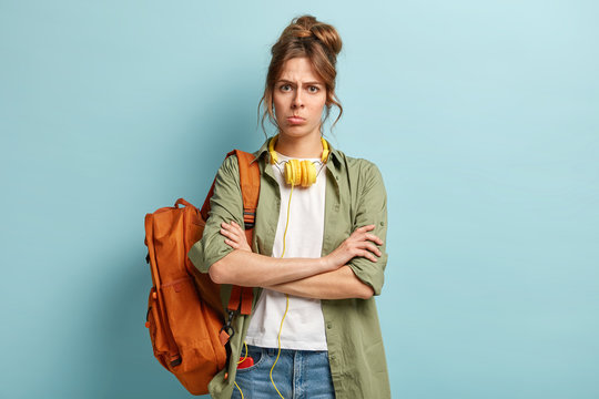 Dissatisfied female keeps arms folded, looks angrily at camera, doesnt want to have classes, carries rucksack, listens audio track in headphones, has dissatisfied facial expression, stands indoor.