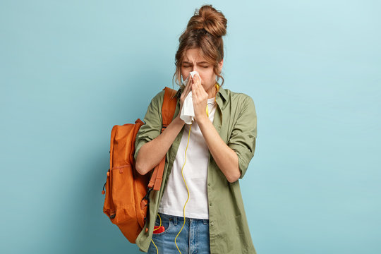 People, disease concept. Ill dark haired woman sneezes in handkerchief, carries rucksack, listens music in headphones, dressed casually, stands against blue background. Student feels unwell.