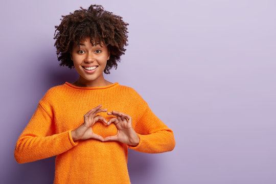 Affectionale lovely woman has heart full of love, shows valentine sign, dressed in casual orange jumper, expresses love, confesses in sympathy, stands in studio against purple wall with blank space