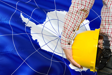 Obraz premium Engineer is holding yellow safety helmet with waving Antarctica flag background. Construction and building concept.