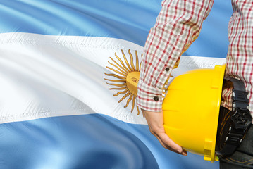 Argentine Engineer is holding yellow safety helmet with waving Argentina flag background. Construction and building concept.