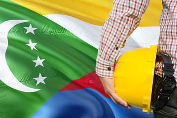 Comoran Engineer is holding yellow safety helmet with waving Comoros flag background. Construction and building concept.
