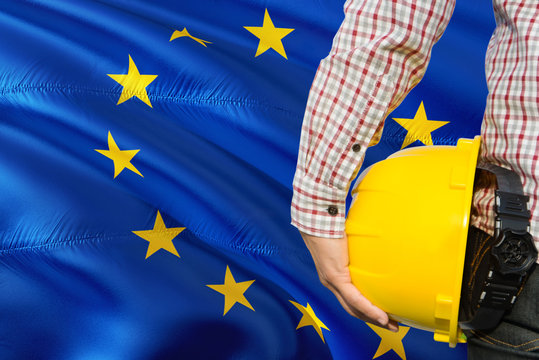 Engineer is holding yellow safety helmet with waving European Union flag background. Construction and building concept.