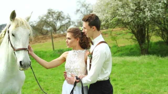 Bride with groom holding white horse and caressing her head to side of them and making wedding teepee making background in nature.