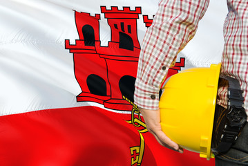 Engineer is holding yellow safety helmet with waving Gibraltar flag background. Construction and...