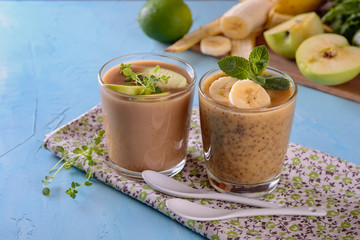 Healthy and tasty yogurt detox smoothies with chia seeds, greenery, bananas, apples and other green ingredients and pieces of ripe bananas in a glasses on a napkin and blue background next to  ingredi