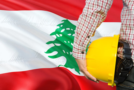 Lebanese Engineer is holding yellow safety helmet with waving Lebanon flag background. Construction and building concept.
