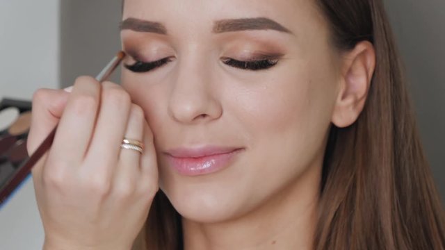 Closeup shot of cheerful young female makeup artist enjoying working with a beautiful young brown haired model, applying fresh trendy makeup on her eyes with long eyelashes, perfect brows using pencil