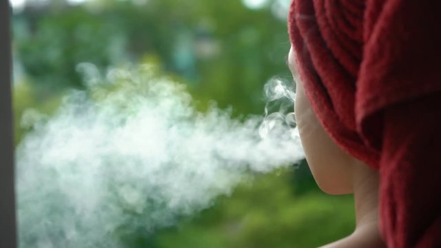 young beautiful caucasian woman with a red towel on her head vaping on the balcony after shower. Close up