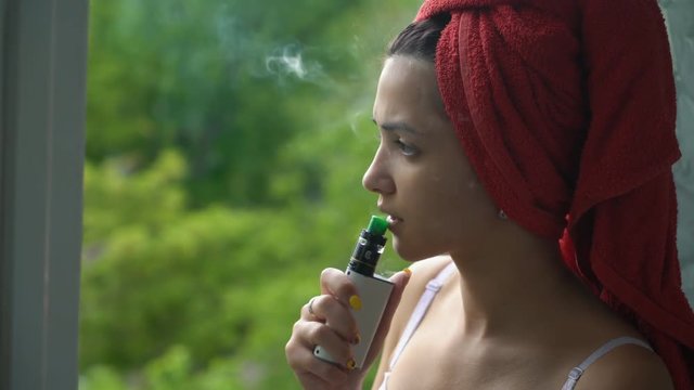 young beautiful caucasian woman in the bra and a red towel on her head vaping on the balcony after shower. Close up
