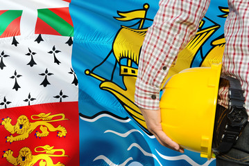 Engineer is holding yellow safety helmet with waving Saint Pierre And Miquelon flag background. Construction and building concept.