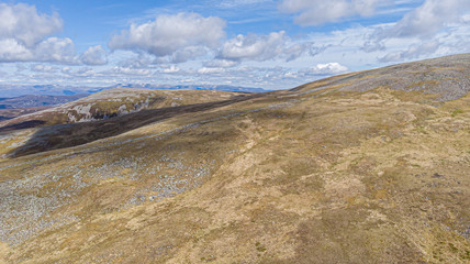An aerial view of a Scottish moountain rocky slope with mountain range in the background under a majestic blue sky and white clouds