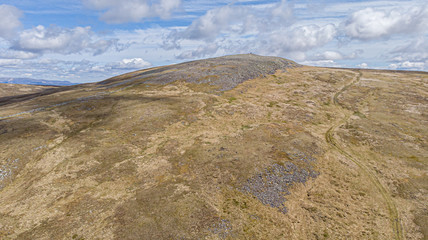 An aerial view of a Scottish rocky Munro summit with trail path under a majestic blue sky and white clouds