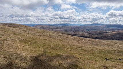 An aerial view of a Scottish moountain green heather slope with mountain range in the background under a majestic blue sky and white clouds