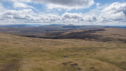 An aerial view of a Scottish mountain summit plateau with heather and trail path  under a majestic blue sky and huge white clouds