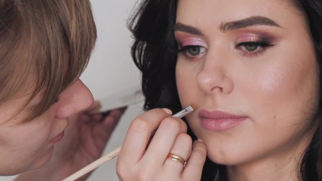 Closeup shot of cheerful young female makeup artist enjoying working with a beautiful young brown haired model, applying fresh trendy makeup on her lips with long eyelashes, perfect brows using pencil