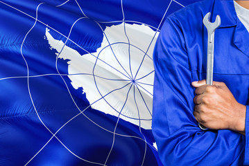 Mechanic in blue uniform is holding wrench against waving Antarctica flag background. Crossed arms technician.