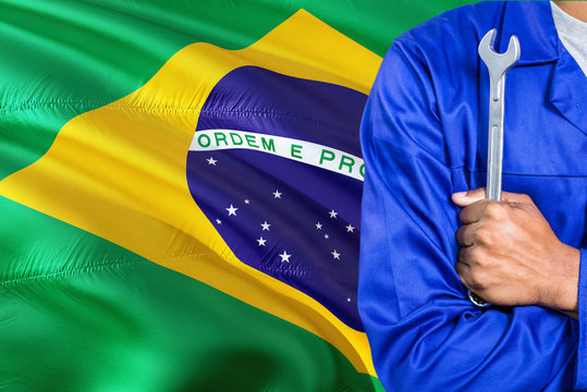 Brazilian Mechanic in blue uniform is holding wrench against waving Brazil flag background. Crossed arms technician.