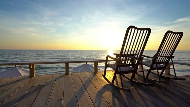 Locked off shot, two rocking chairs on wooden deck, golden sunset sea view background.
