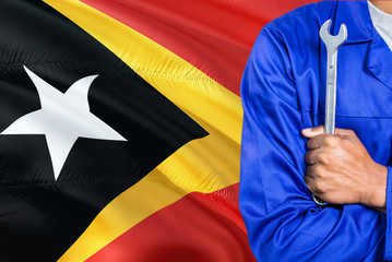 Timorese Mechanic in blue uniform is holding wrench against waving East Timor flag background. Crossed arms technician.