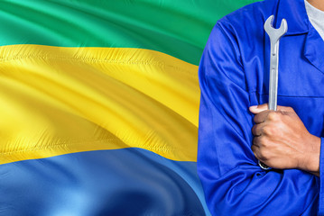 Gabonese Mechanic in blue uniform is holding wrench against waving Gabon flag background. Crossed...