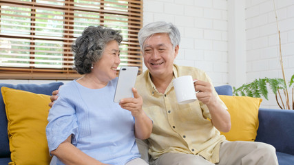 Happy senior asian couple using smart phone while sitting on sofa at home living room background, active senior people retirement and technology lifestyles