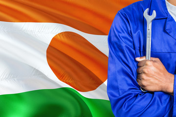 Nigerien Mechanic in blue uniform is holding wrench against waving Niger flag background. Crossed arms technician.