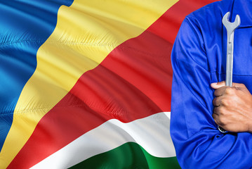 Seychellois Mechanic in blue uniform is holding wrench against waving Seychelles flag background. Crossed arms technician.