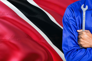 Mechanic in blue uniform is holding wrench against waving Trinidad And Tobago flag background. Crossed arms technician.
