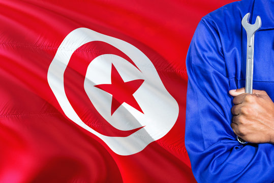 Tunisian Mechanic in blue uniform is holding wrench against waving Tunisia flag background. Crossed arms technician.