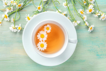 A cup of chamomile tea, shot from above on a teal blue background with fresh flowers and a place...