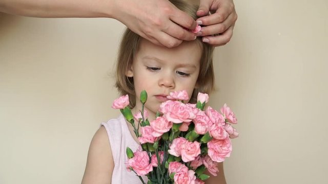 Portrait of little caucasian cute smiling girl stands and holds bouquet of pink carnations, mother's hands pins flower in daughter's hair. Mother's love and care