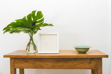 Oak side table with monstera leaves in vase, green bowl and blank square picture frame  against white wall © Natalie Board