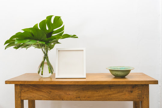 Oak side table with monstera leaves in vase, green bowl and blank square picture frame  against white wall