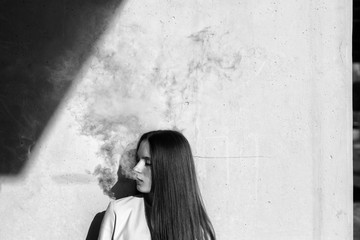 Vape teenager. Young cute girl in  casual clothes smokes an electronic cigarette near the wall outdoors in summer day. Bad habit that is harmful to health. Vaping activity. Black and white. Close up.
