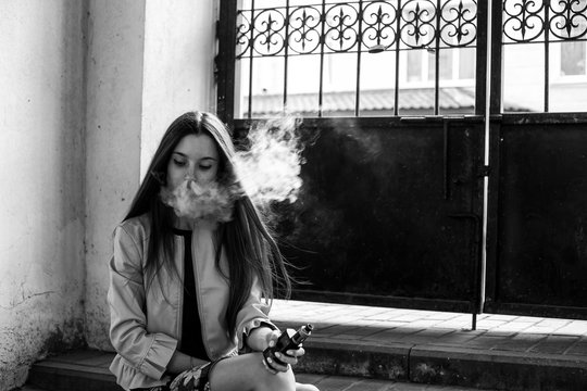 Vape teenager. Young cute girl in casual clothes smokes an electronic cigarette near the wall in prison cell. Bad habit that is harmful to health. Vaping activity. Black and white.
