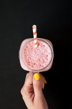 Female hand holding a glass jar filled with strawberry banana smoothie over black background, overhead view. Top view, from above, flat lay. Close-up.