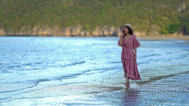 Gorgeous young Asian woman in a pink dress is walking along a beach while talking on her telephone and smiling.