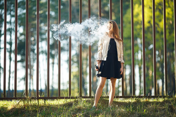 Vape teenager. Young cute girl in casual clothes smokes an electronic cigarette in front of a metal...