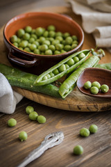 view of some raw fresh green peas in an earthenware plate and some pea pods on a brown rustic wooden table and linen